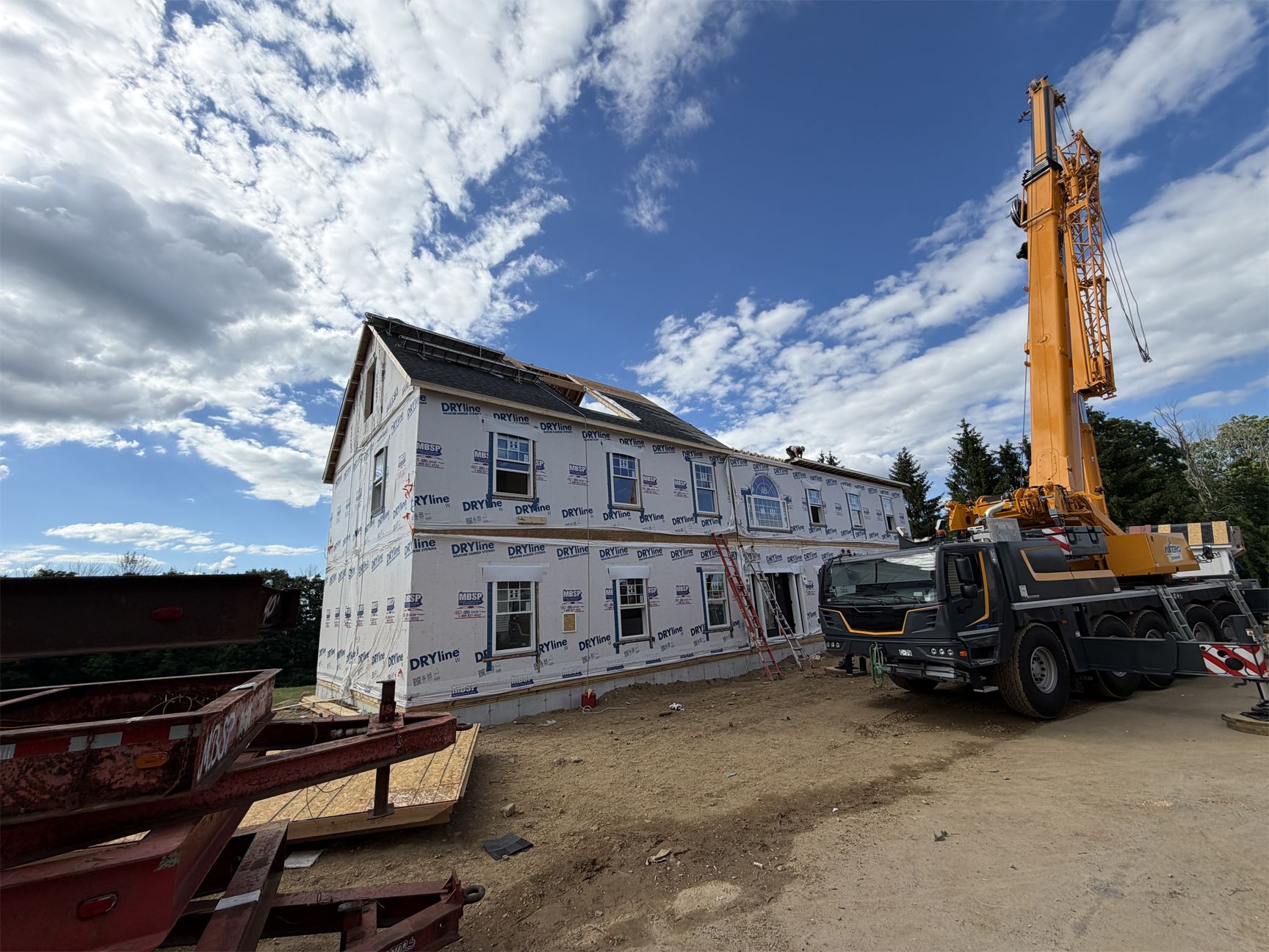 House under construction beside a yellow drilling rig on a gravel site under a cloudy sky