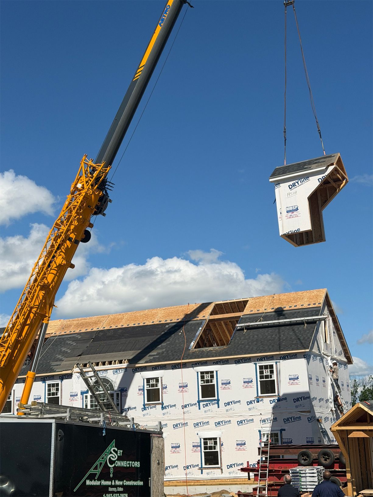 Crane lifting a house section over a partially rebuilt white house under a blue sky