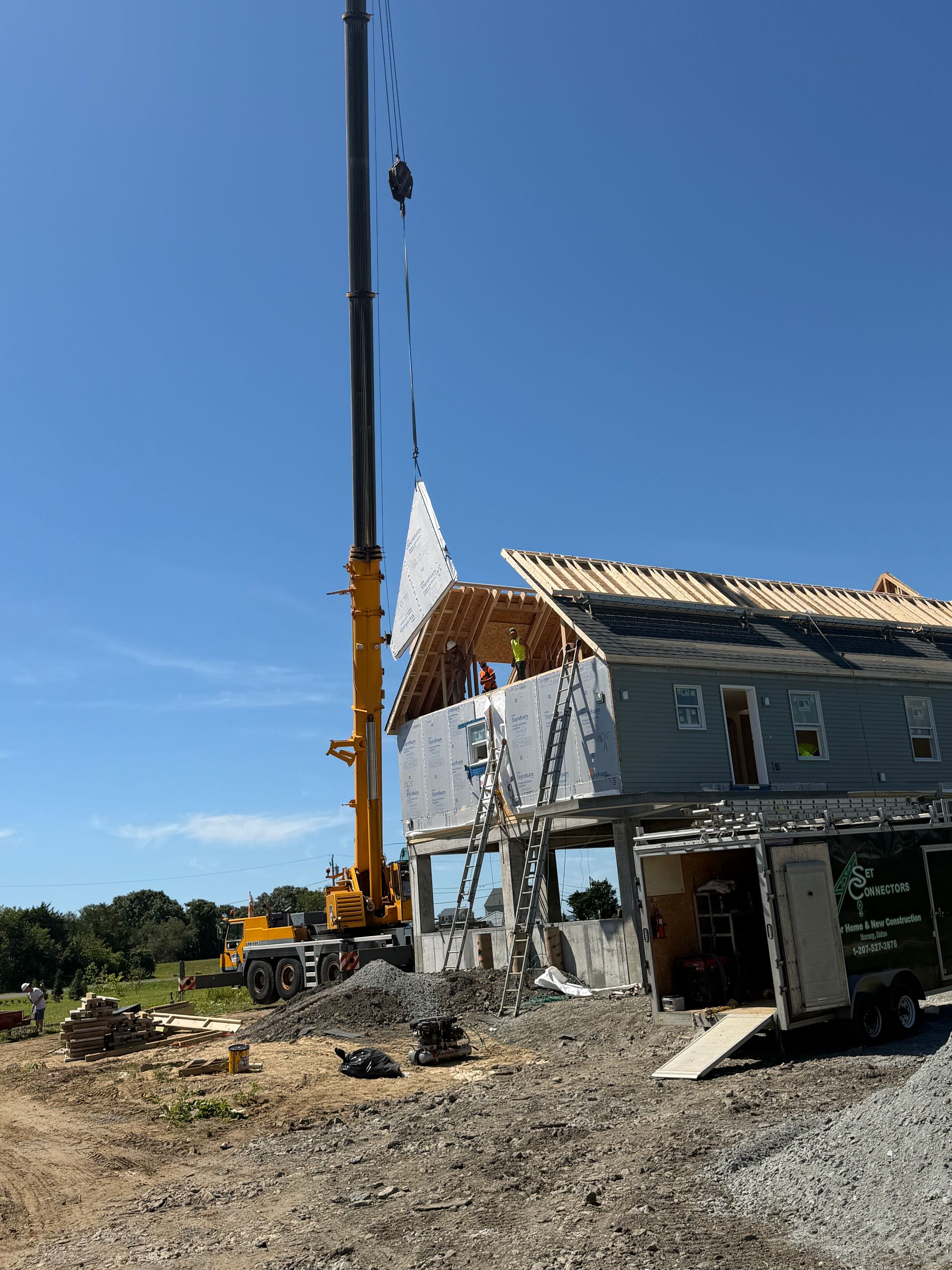 Construction site with crane lifting materials beside a partially built gray house under a blue sky