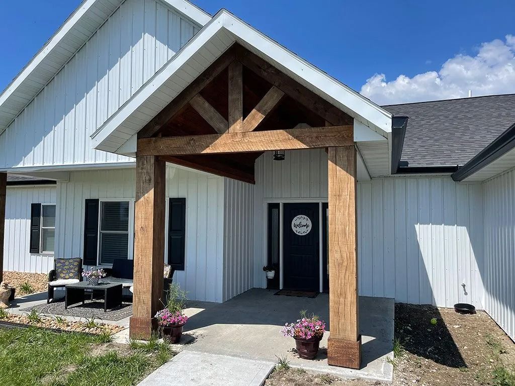 White house with brown wooden porch. Blue door, flowers, black shutters, and a table.