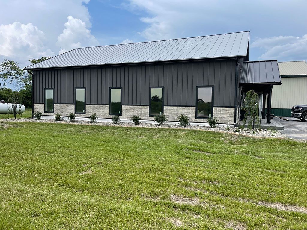 Gray building with metal roof, windows, and stone base. Green grass in the foreground, cloudy sky.