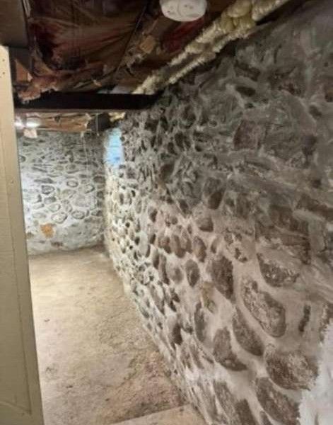 Stone basement interior with a dirt floor, showing walls, ceiling beams, and a light fixture.