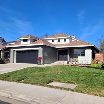 A single-story house with green siding, a black garage door, and a brown roof under a blue sky.