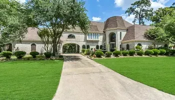 Large beige house with long driveway and well-manicured lawn under a blue sky with trees.