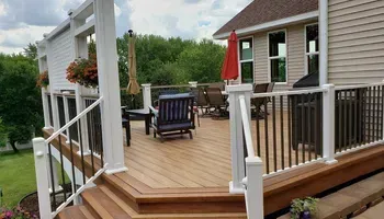 Wooden deck with seating and an umbrella, overlooking a green landscape, attached to a light-colored house.