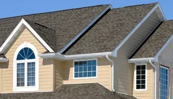 Tan house with dark gray shingled roof, white trim, and blue windows under a sunny sky.