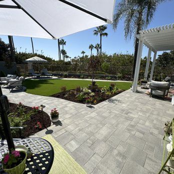 Patio with gray pavers, green lawn, flower beds, umbrella, pergola, palm trees. Sunny day.