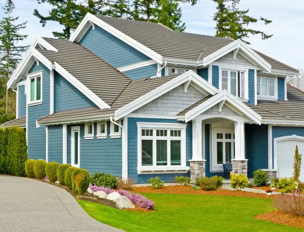 Blue house with white trim, gray roof, and green lawn. Driveway leads up to the front.