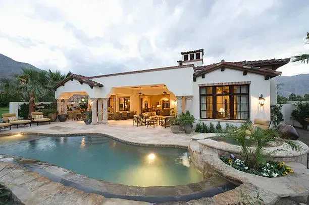 Poolside view of a white stucco house with a tiled roof, pool, and patio furniture.