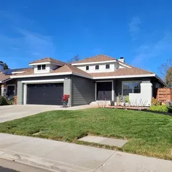 A single-story house with green siding, a black garage door, and a brown roof under a blue sky.