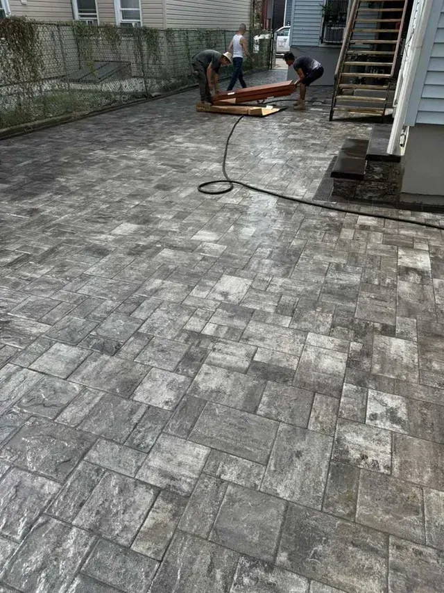 Workers installing paving stones in a gray, patterned driveway. One person holds wooden beams.