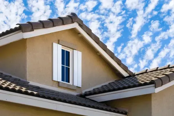 Tan house with brown tile roof, white window shutters, blue sky with clouds.