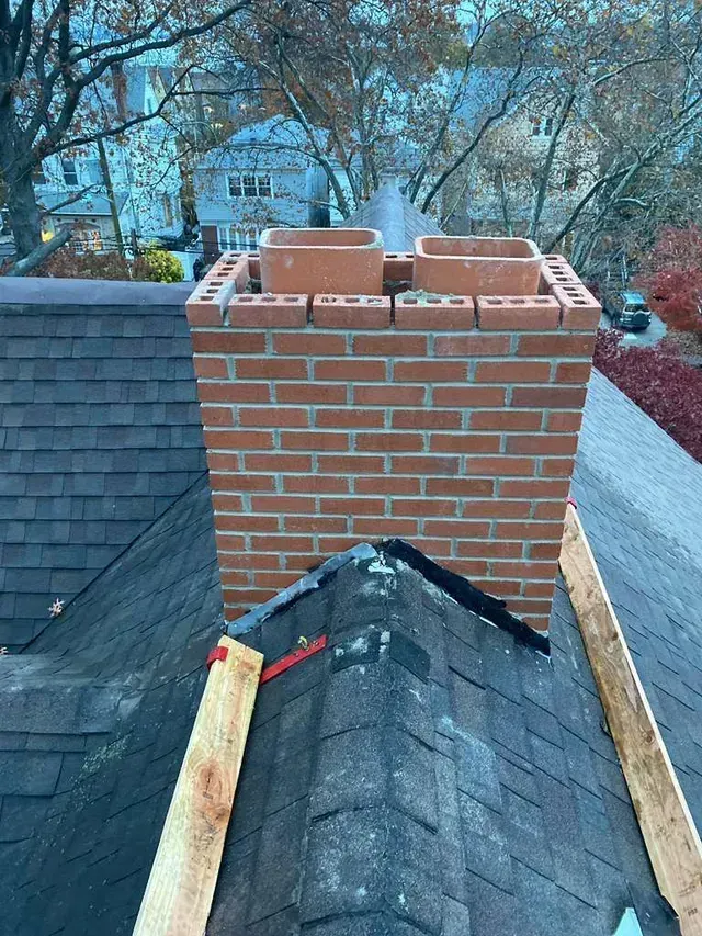 Brick chimney on a dark shingle roof with two clay flue liners; wooden boards on roof edges.
