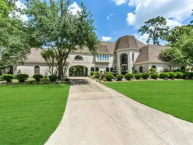 A large, light-colored mansion with a long driveway and well-kept lawn under a blue sky.