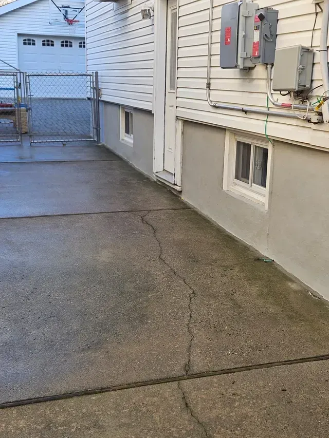 Concrete patio next to a house with a door, windows, and electrical boxes. A chain-link fence is visible.