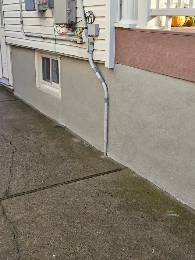 Exterior wall with window and conduit, next to a concrete walkway.