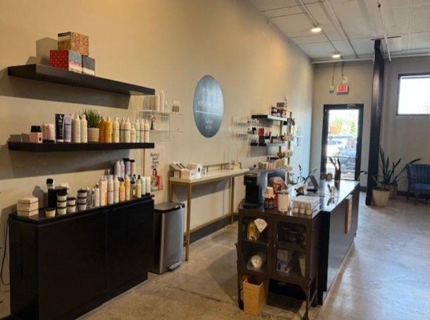 Bright salon interior with black shelves, hair products, styling stations, and a front door visible at the end.