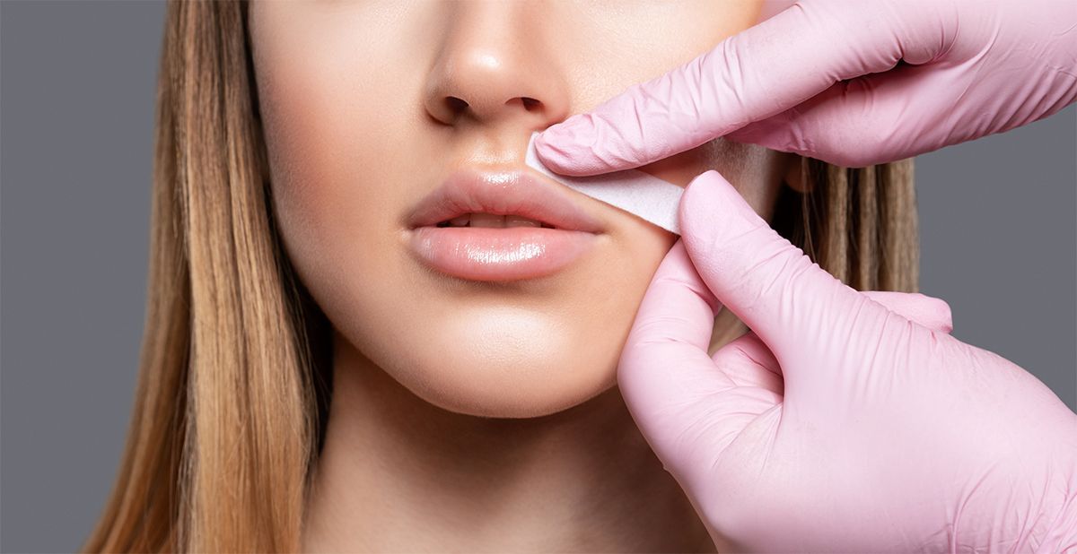 A person in pink gloves uses a wax strip to remove hair from above a person's upper lip.
