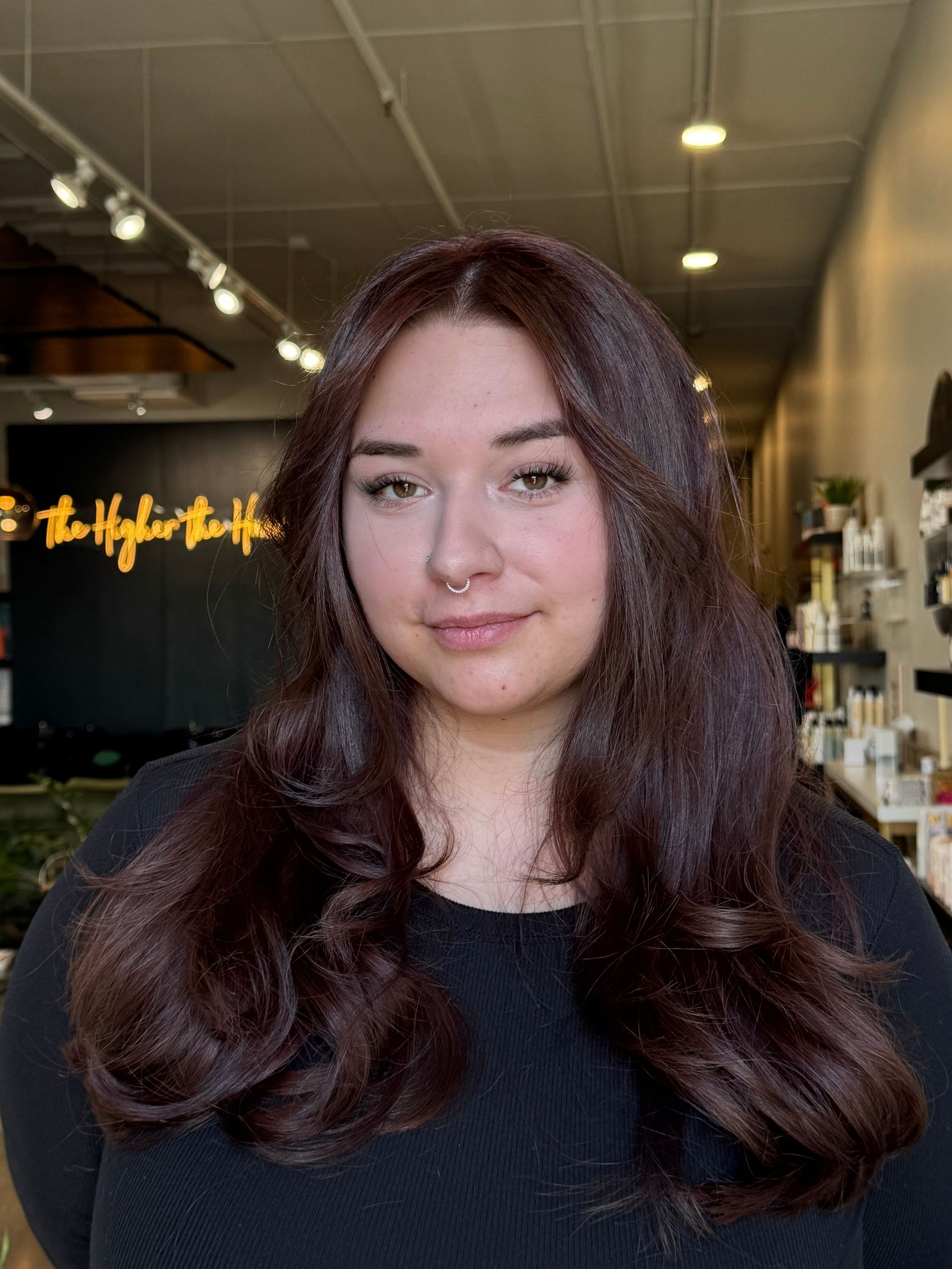 A person with long, dark brown hair and a neutral expression in a brightly lit salon with a black accent wall.