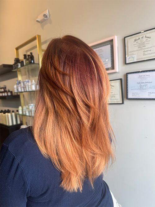 A person with shoulder-length, layered auburn to strawberry blonde gradient hair, seen from behind in a salon.
