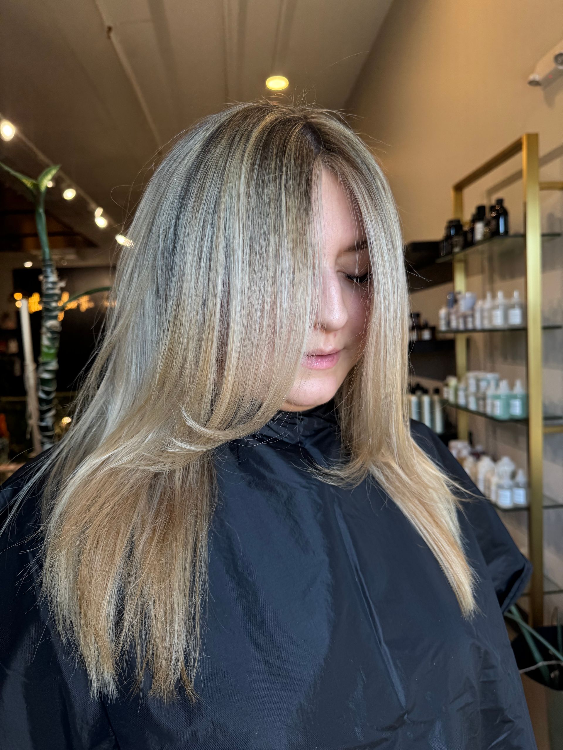 A person with long, textured, blonde-highlighted hair in a salon, wearing a black cape.