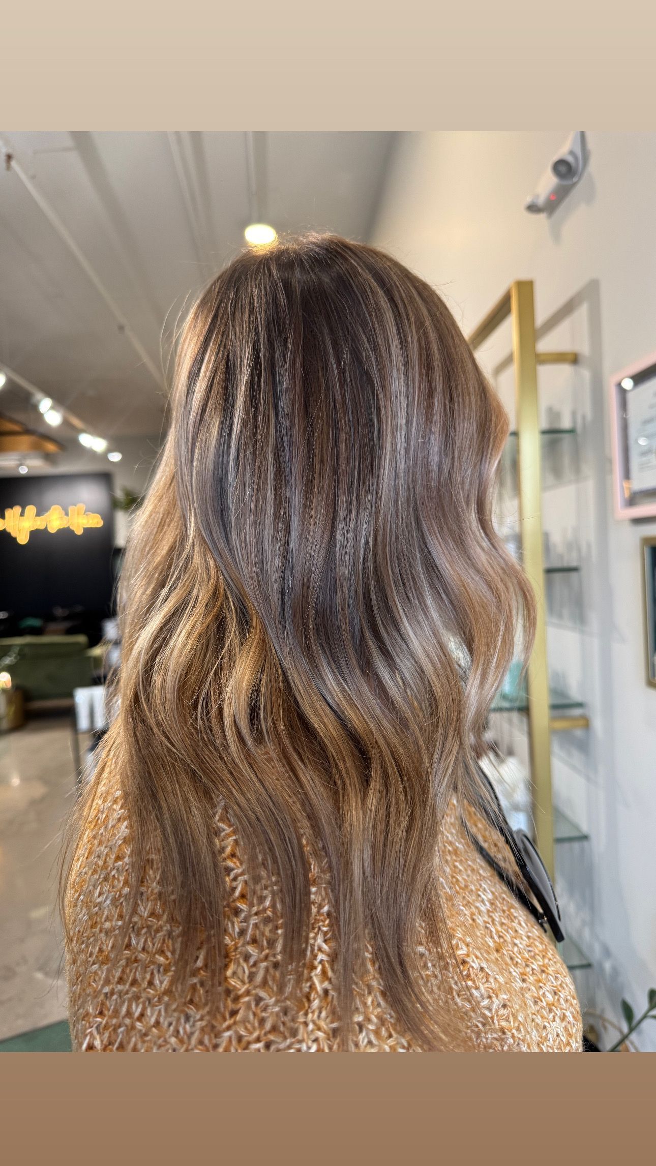 Back view of hair with brown roots transitioning into lighter, caramel-toned highlights in a salon setting.