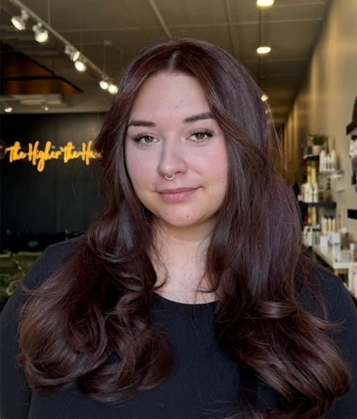 A person with long, dark brown, wavy hair and a septum piercing, looking at the camera in a hair salon.