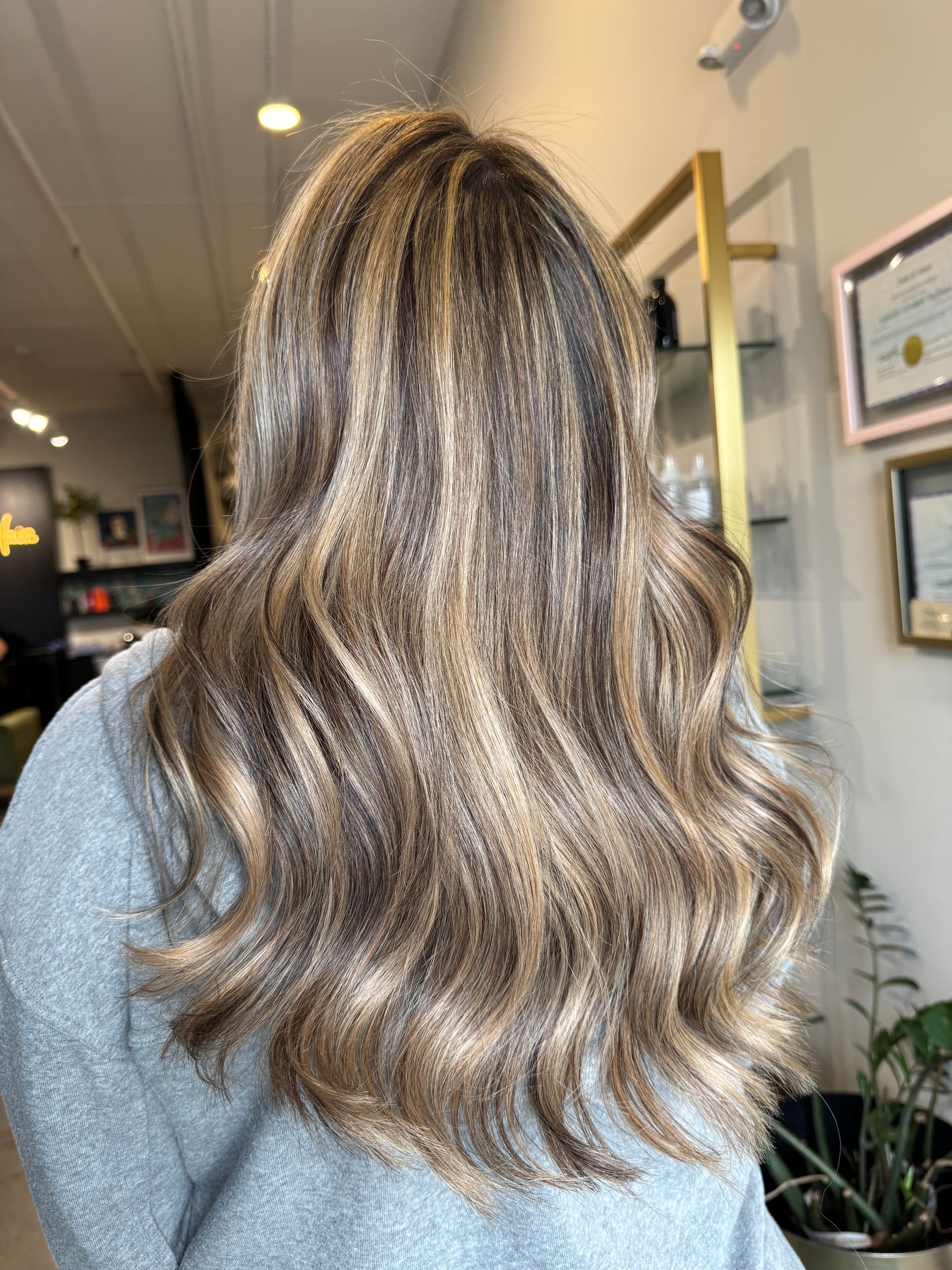 A person with long, wavy, highlighted brunette hair styled in a salon, viewed from the back.