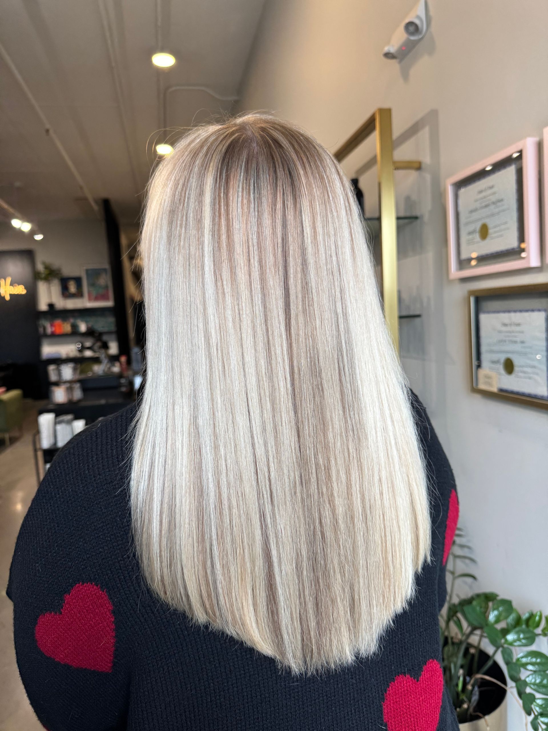 Back view of a person with straight, multi-tonal blonde hair in a salon setting, wearing a black sweater with red hearts.