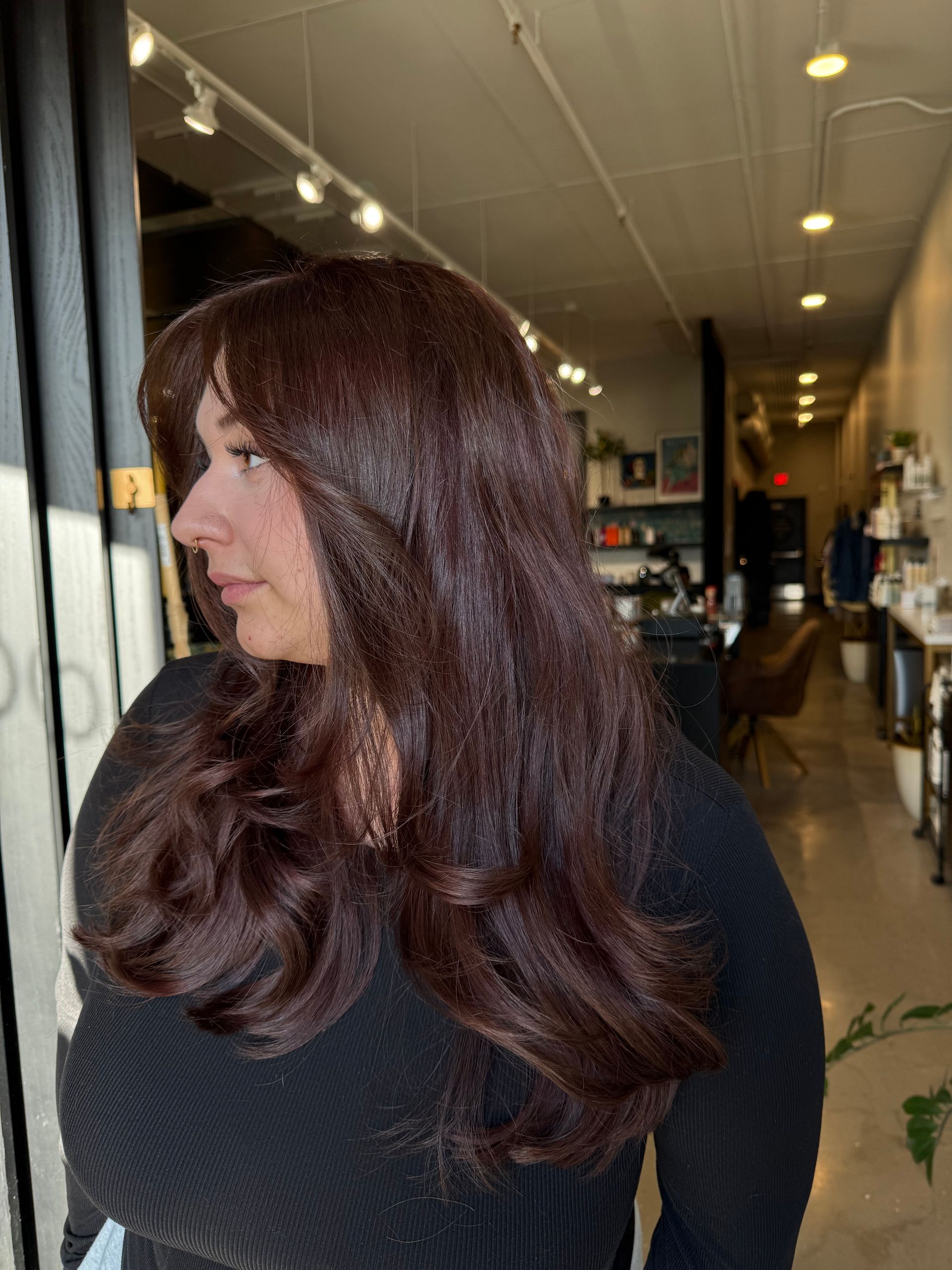 A person with long, voluminous, deep auburn-brown hair styled in soft layers, viewed in profile inside a hair salon.