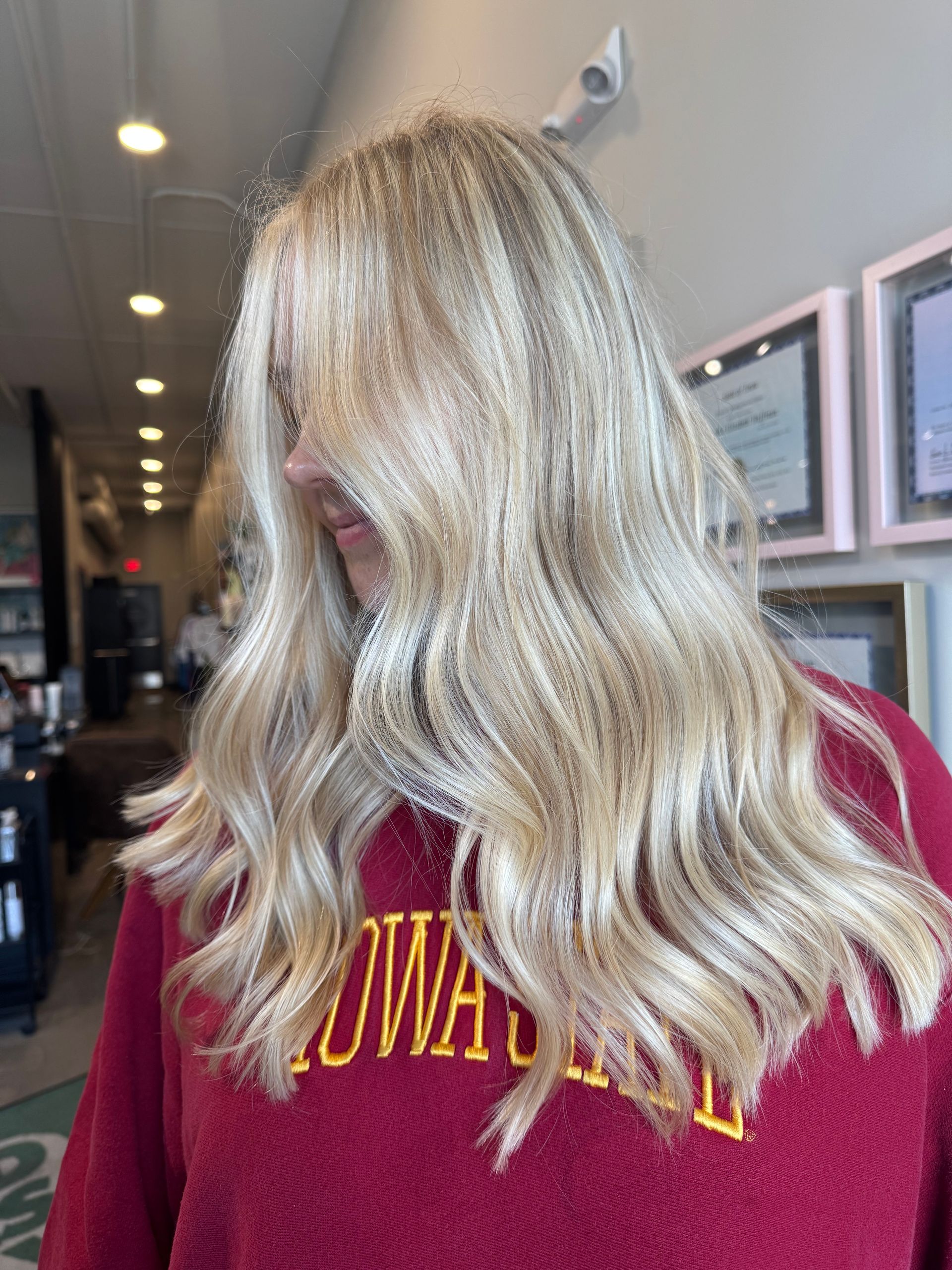 A person with long, wavy platinum blonde hair wears a red sweatshirt in a salon setting.