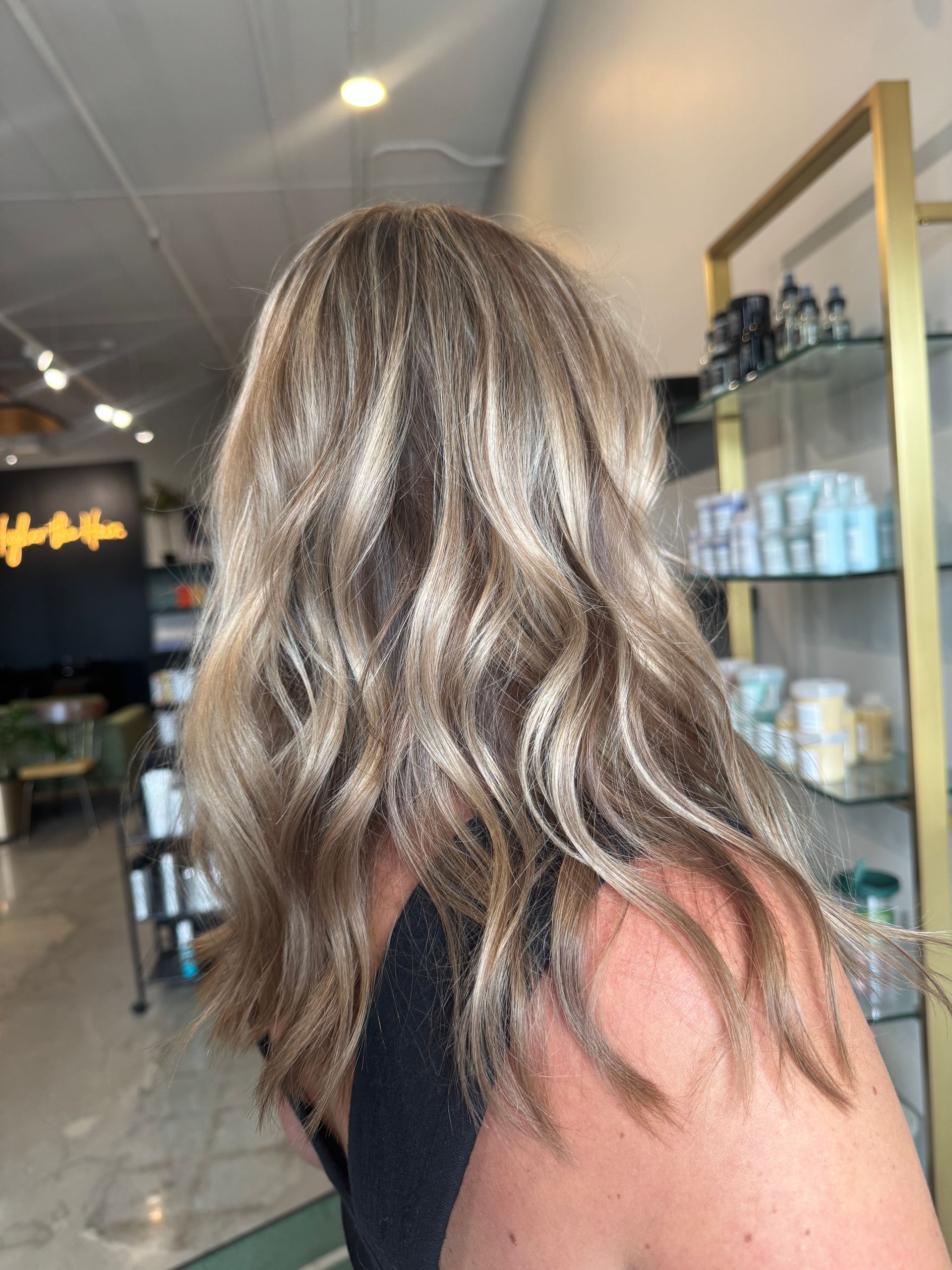 A person with wavy, highlighted blonde hair viewed from behind, standing inside a hair salon.