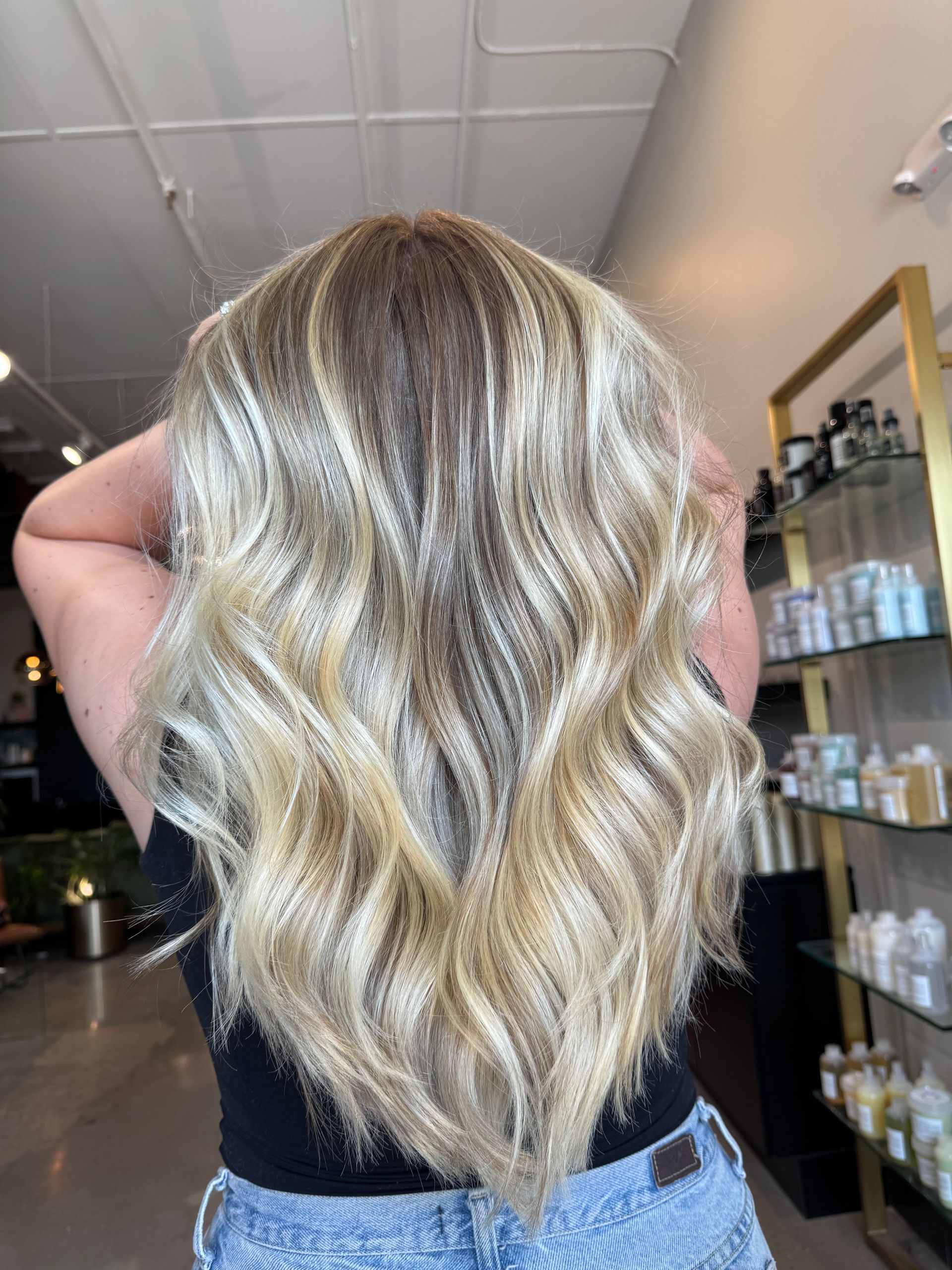 A person from behind displays long, wavy, highlighted blonde hair with dark roots in a salon setting.