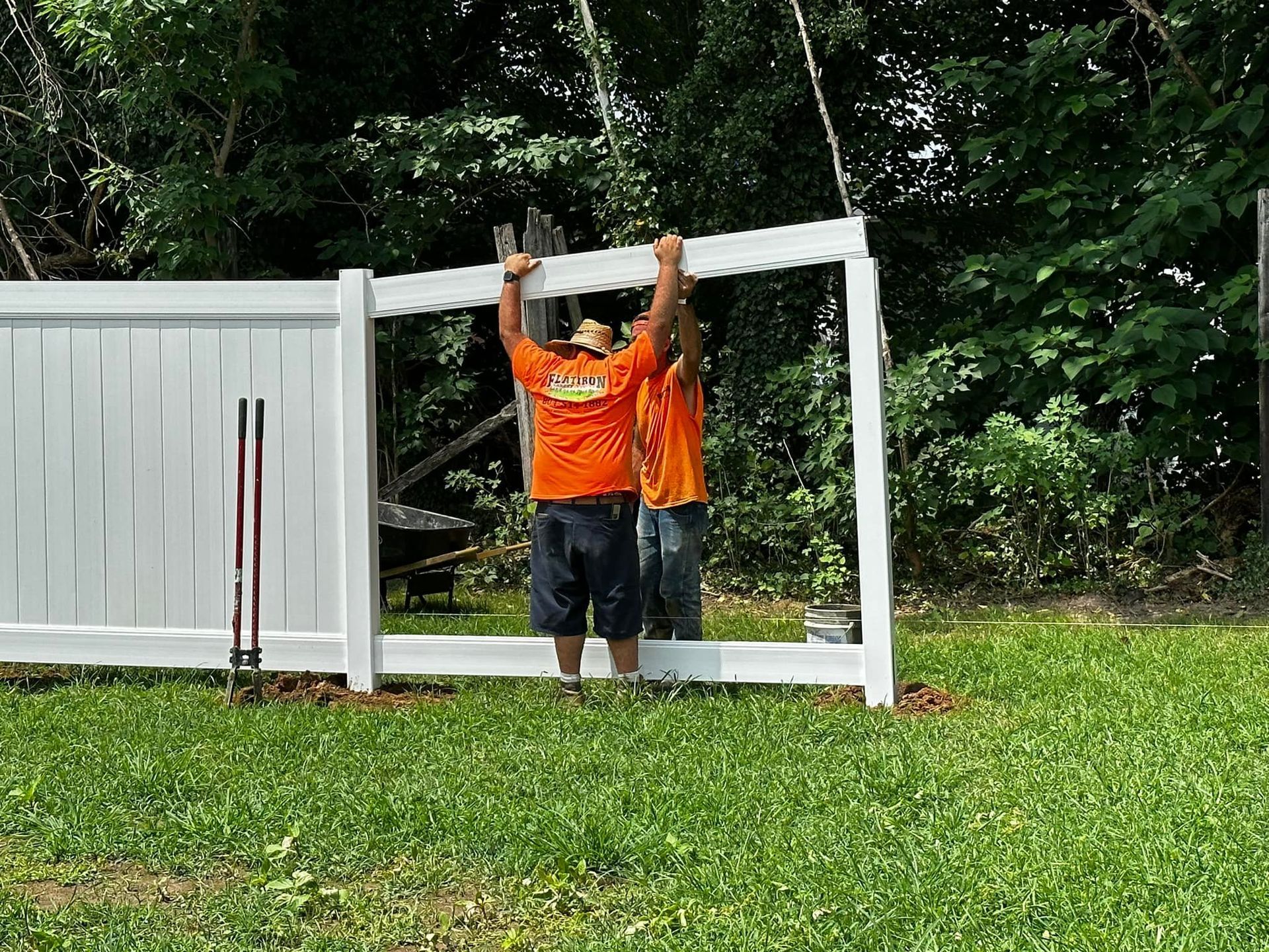 Two men in orange shirts are installing a white fence.