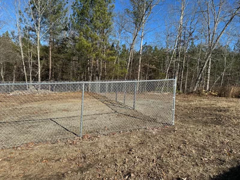 A chain link fence surrounds a dirt field with trees in the background.