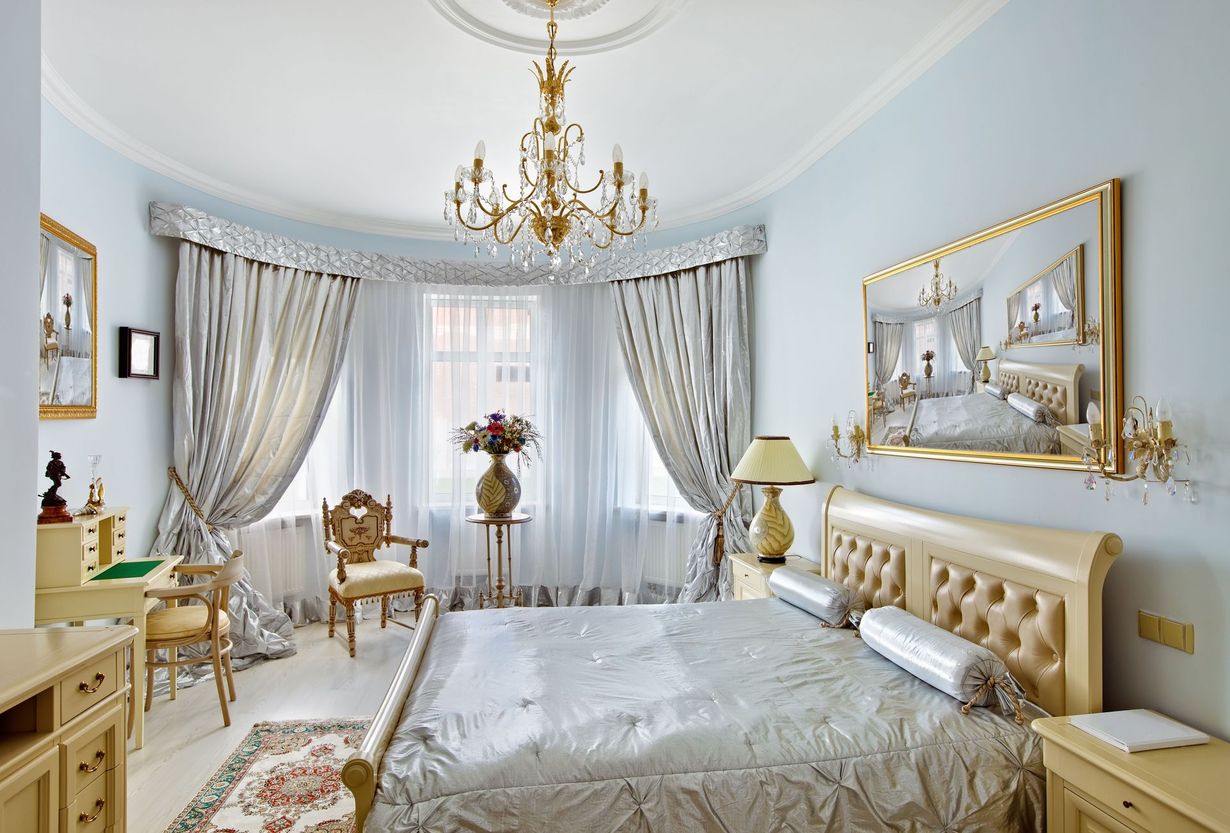 Elegant light-blue bedroom with gold accents, chandelier, tufted bed, and large draped windows