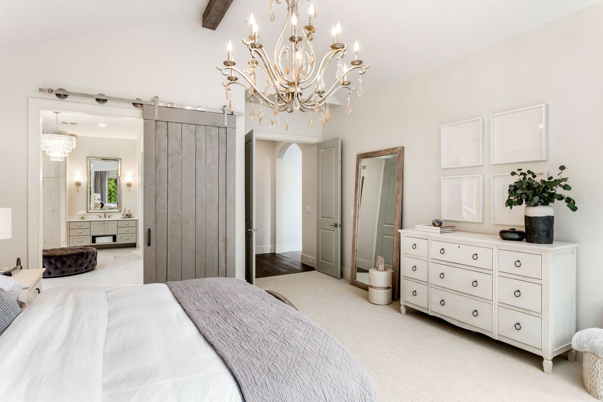 Bright bedroom with a chandelier, bed, white dresser, and leaning mirror in a neutral-toned room.