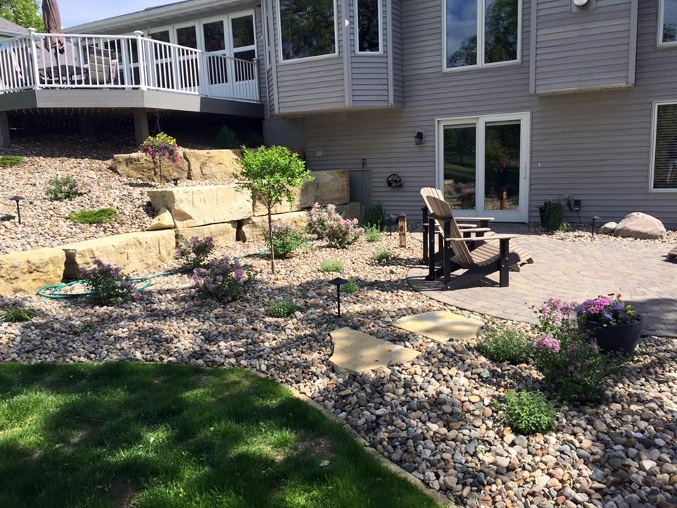 Backyard with retaining wall, patio, and Adirondack chair. Gravel, plants, deck, and house. Sunny day.