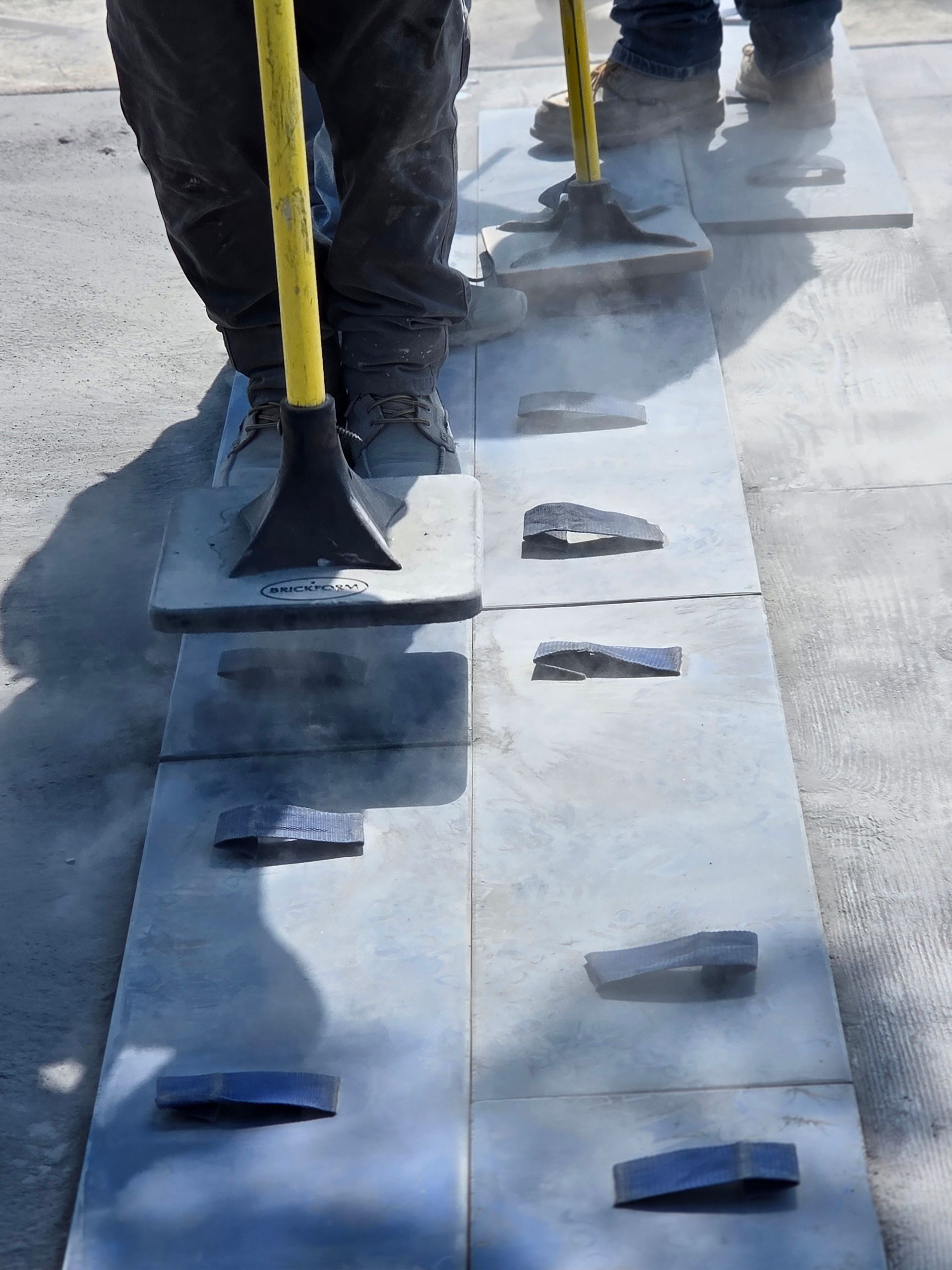Workers use tools to install rectangular pavement stones, producing dust.
