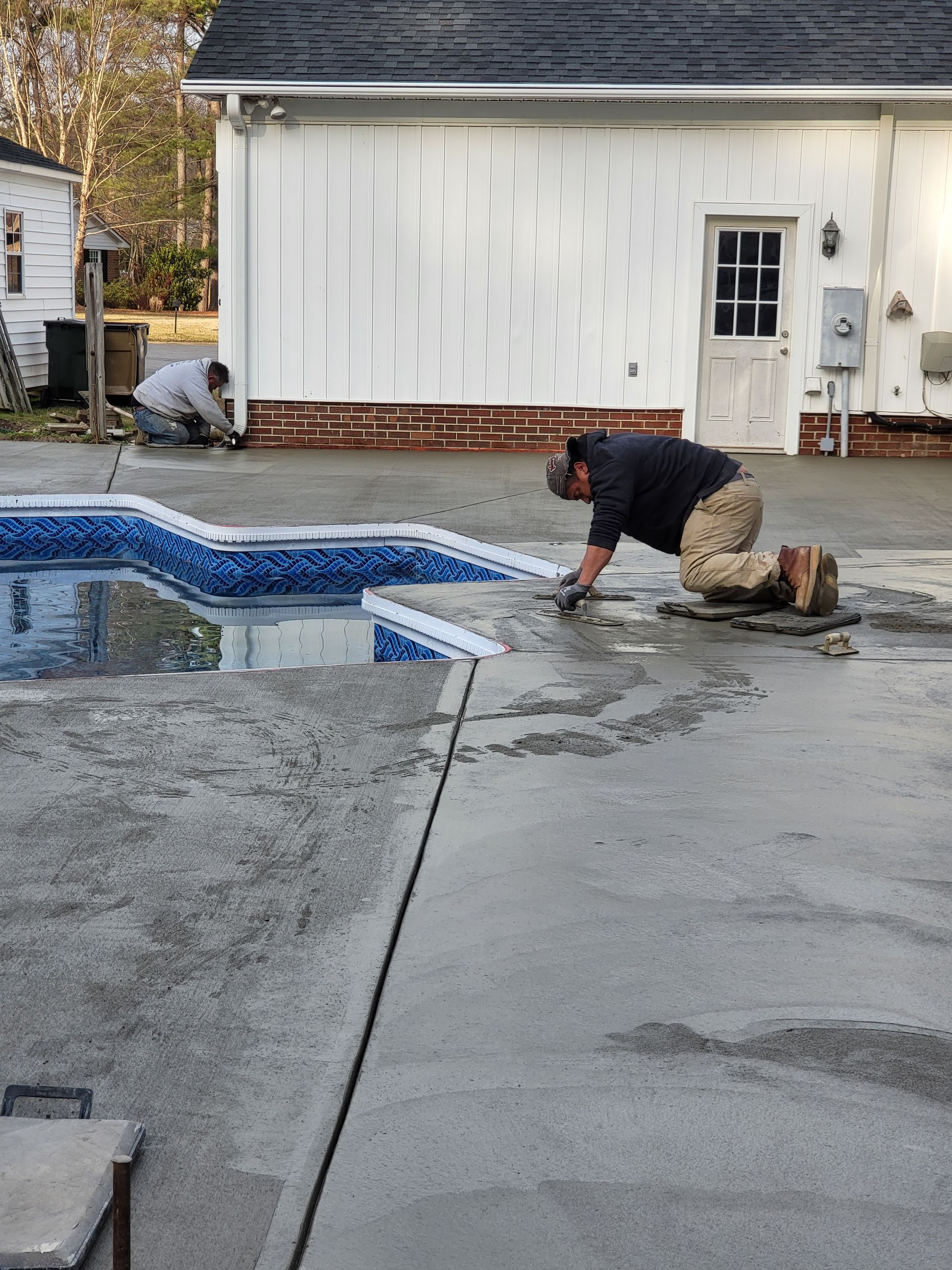 Two people work on concrete around a pool. One kneels and applies material, another works in the background. White building in the background.