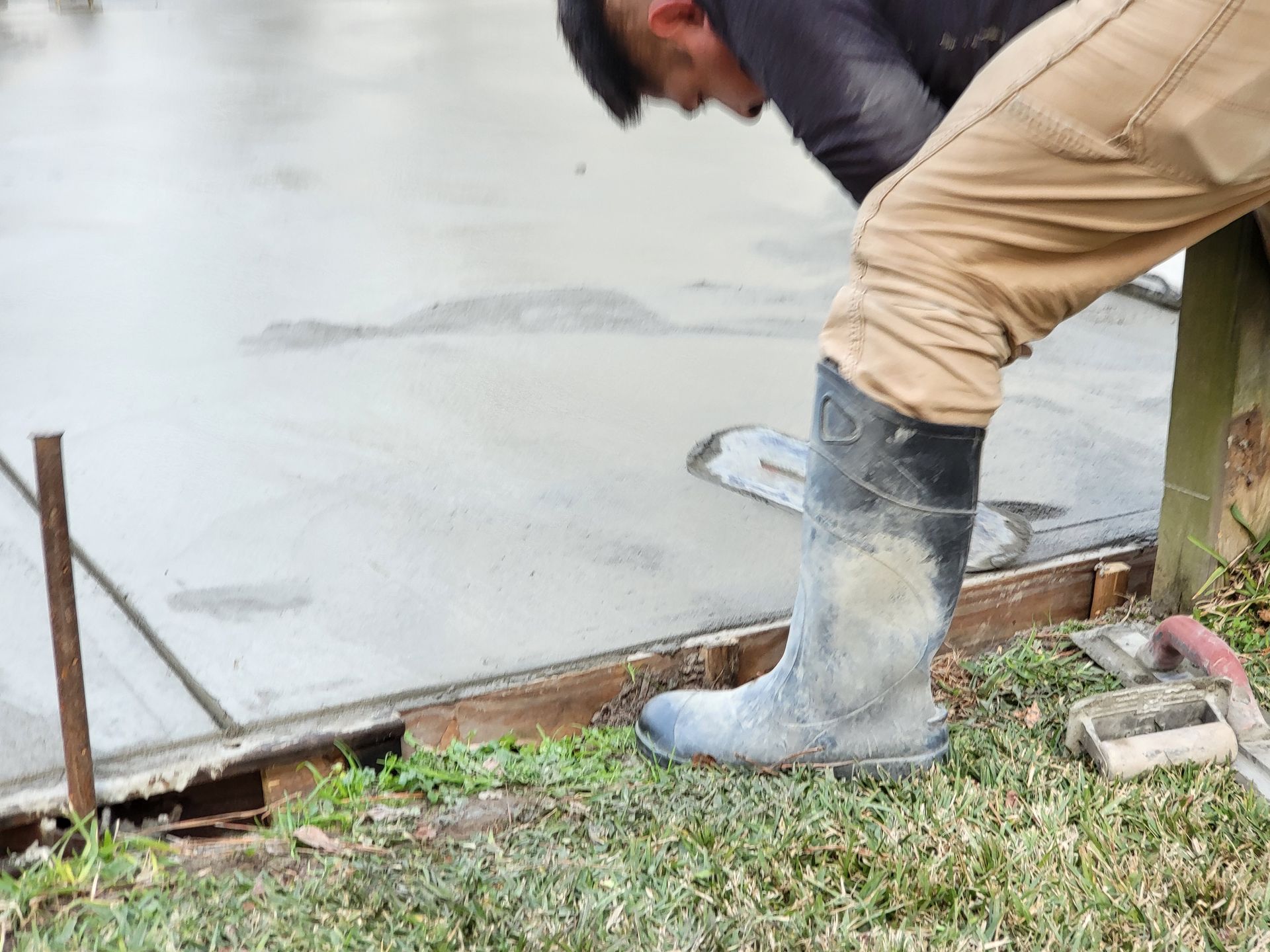 Person smoothing wet concrete with a trowel, wearing rubber boots and khaki pants, outdoors.