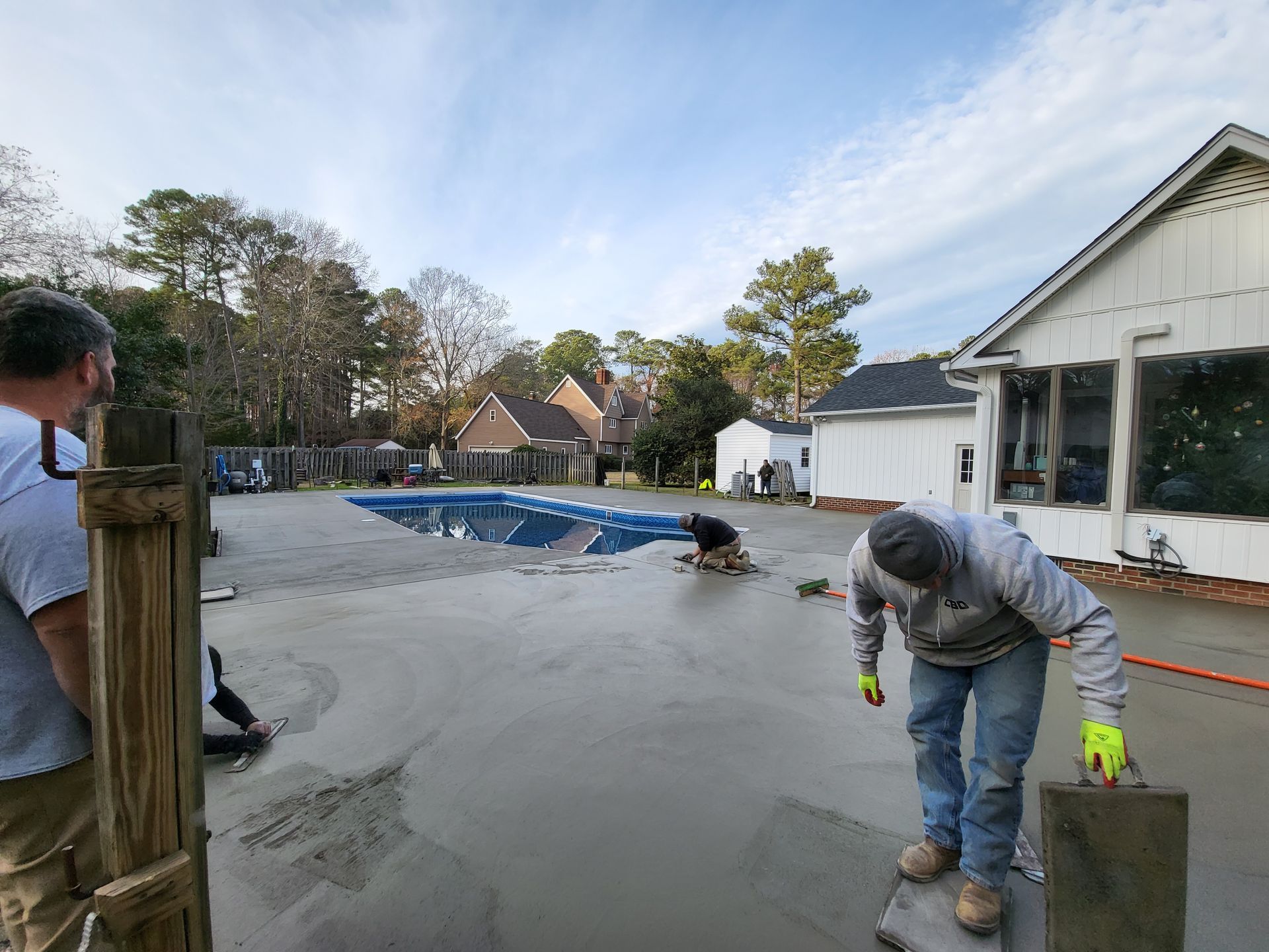 Workers smoothing wet concrete around a swimming pool; residential setting with a sunny sky.