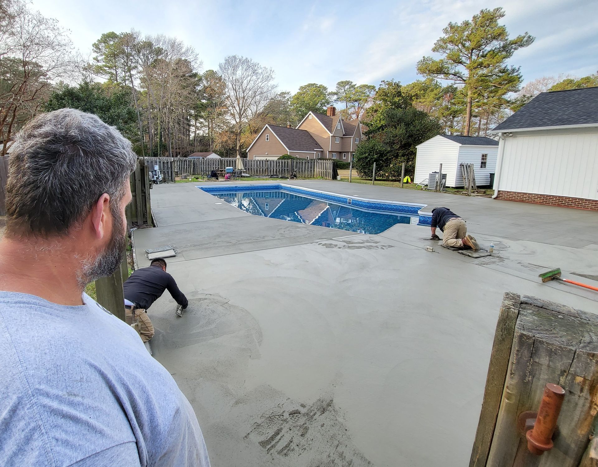 Workers pouring concrete around a pool; one man observes. Outdoor setting with houses and trees.