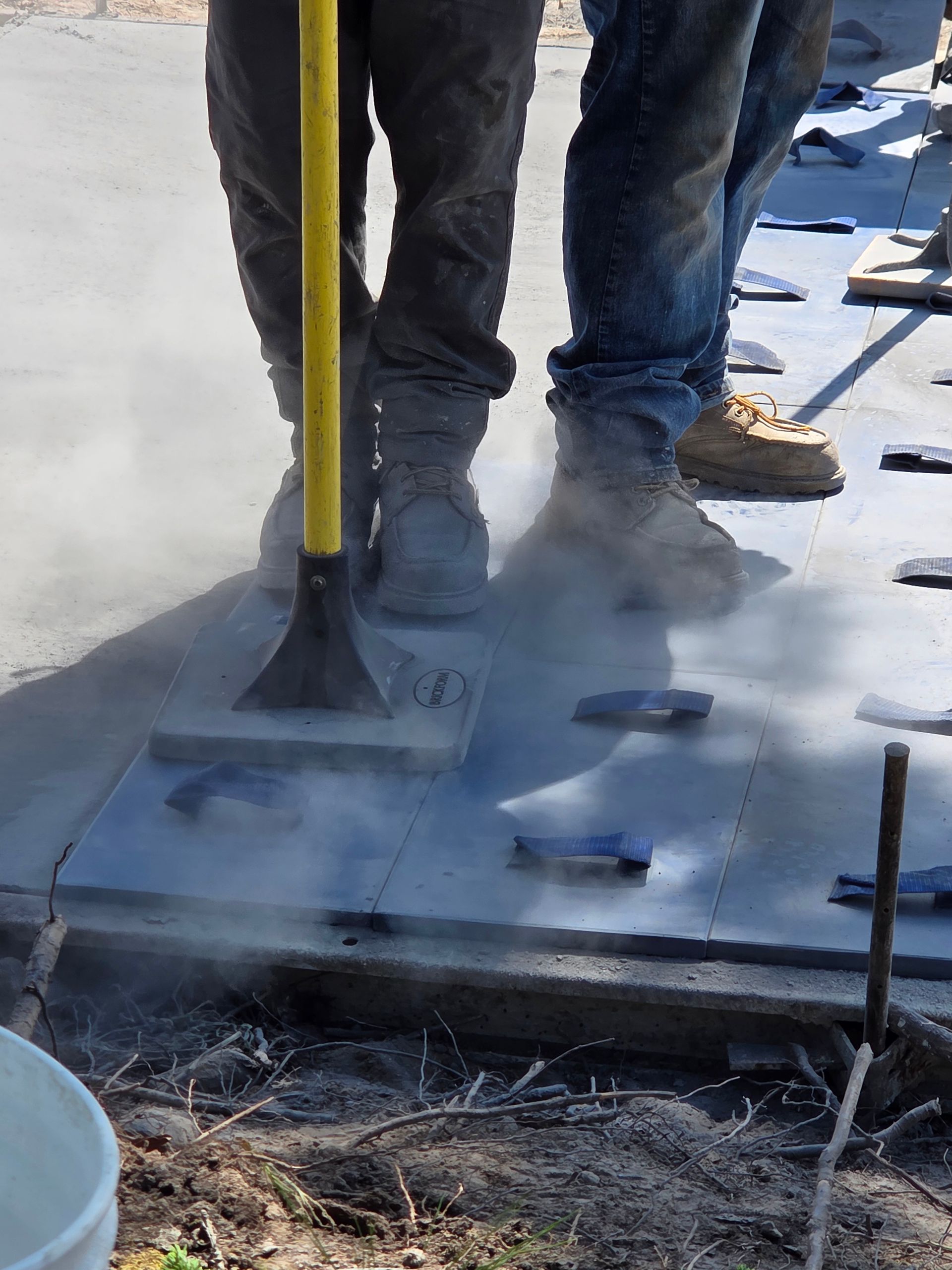Two workers compacting material with a power compactor on a construction site; visible dust.