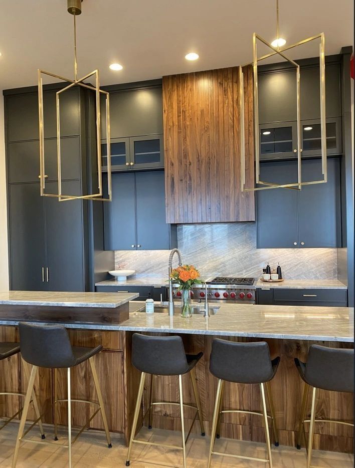 Modern kitchen with dark cabinets, wooden range hood, gold light fixtures, and bar stools.