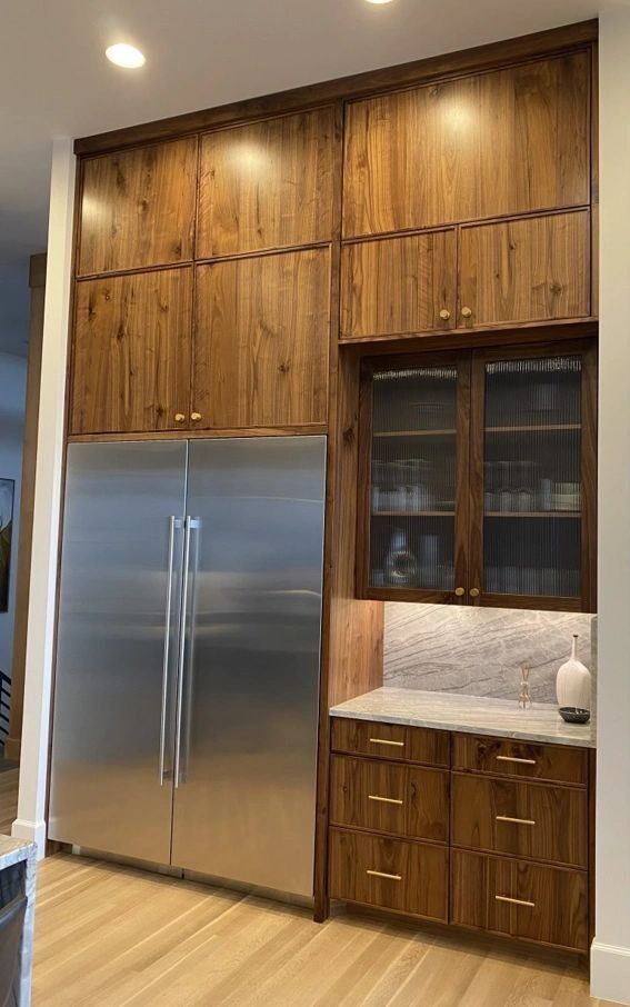 Tall wood cabinets surround a stainless steel refrigerator, with a countertop and glass-front cabinet.
