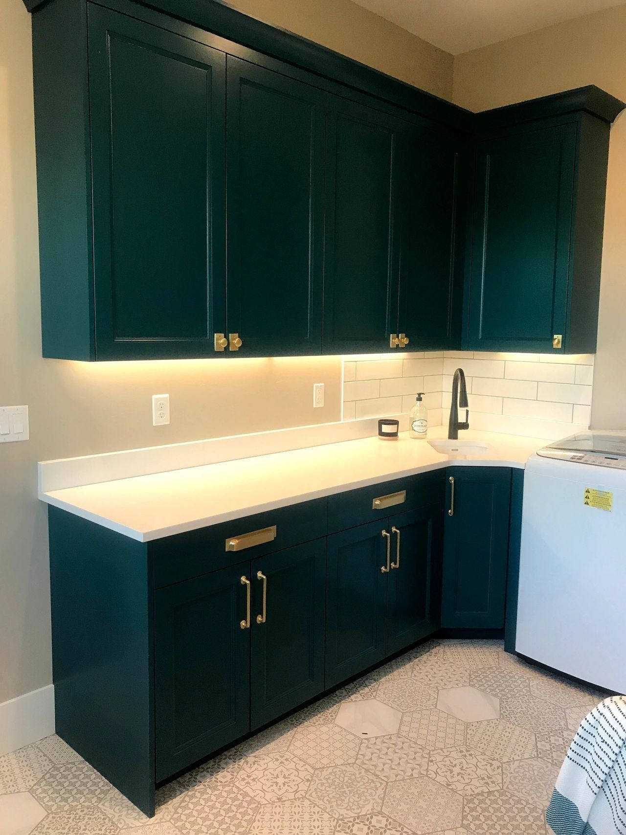 Dark teal cabinets with gold hardware in a laundry room. Beige countertop, tiled backsplash, and patterned floor.