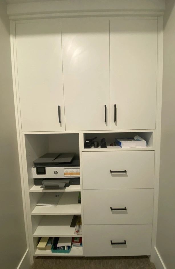 White built-in cabinet with upper doors, center open shelf/drawer section, and lower drawers; holds printer and supplies.