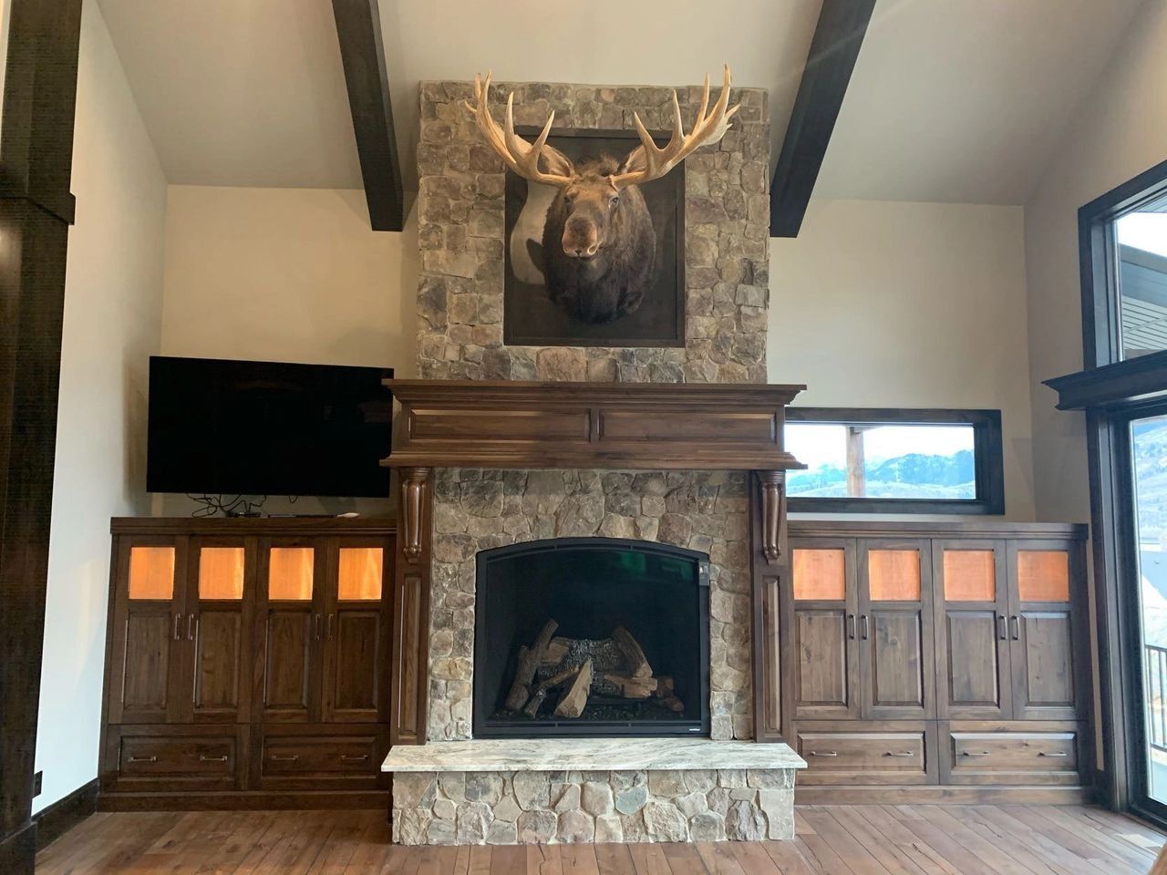 Living room with stone fireplace, moose head, TV, and wooden cabinets.
