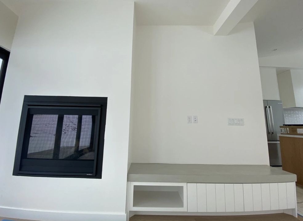 Modern living room with white walls, black fireplace, concrete bench, and kitchen in the background.